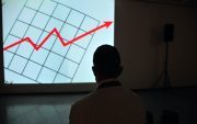 Silhouette of a man staring at a graph on a projector screen. Red arrow moving up and to the right is visible on the screen