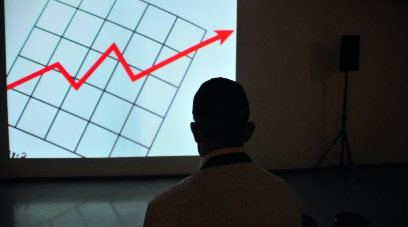 Silhouette of a man staring at a graph on a projector screen. Red arrow moving up and to the right is visible on the screen